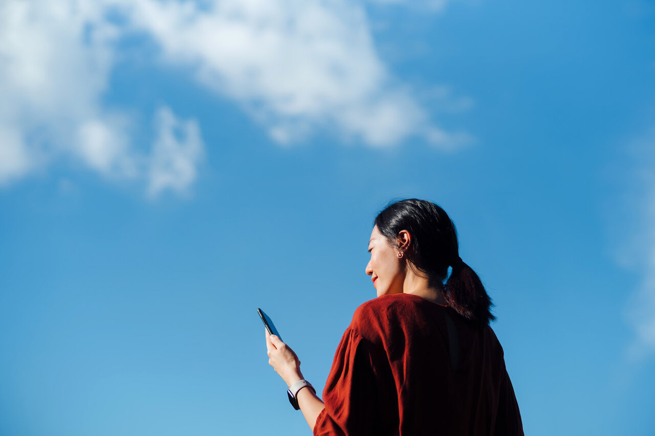 Low angle portrait of young Asian woman using smartphone against beautiful blue sky with cloudscapes, enjoying sunlight outdoors. Lifestyle and technology | © © 2021 Yiu Yu Hoi