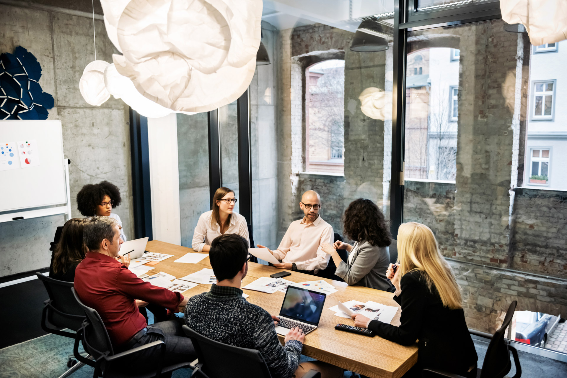 A group of young people, male and female, of different ethnicities are sitting in a bright modern office room. They are discussing something during a business meeting. There are documents and laptops on the table. | © Hinterhaus Productions @Getty