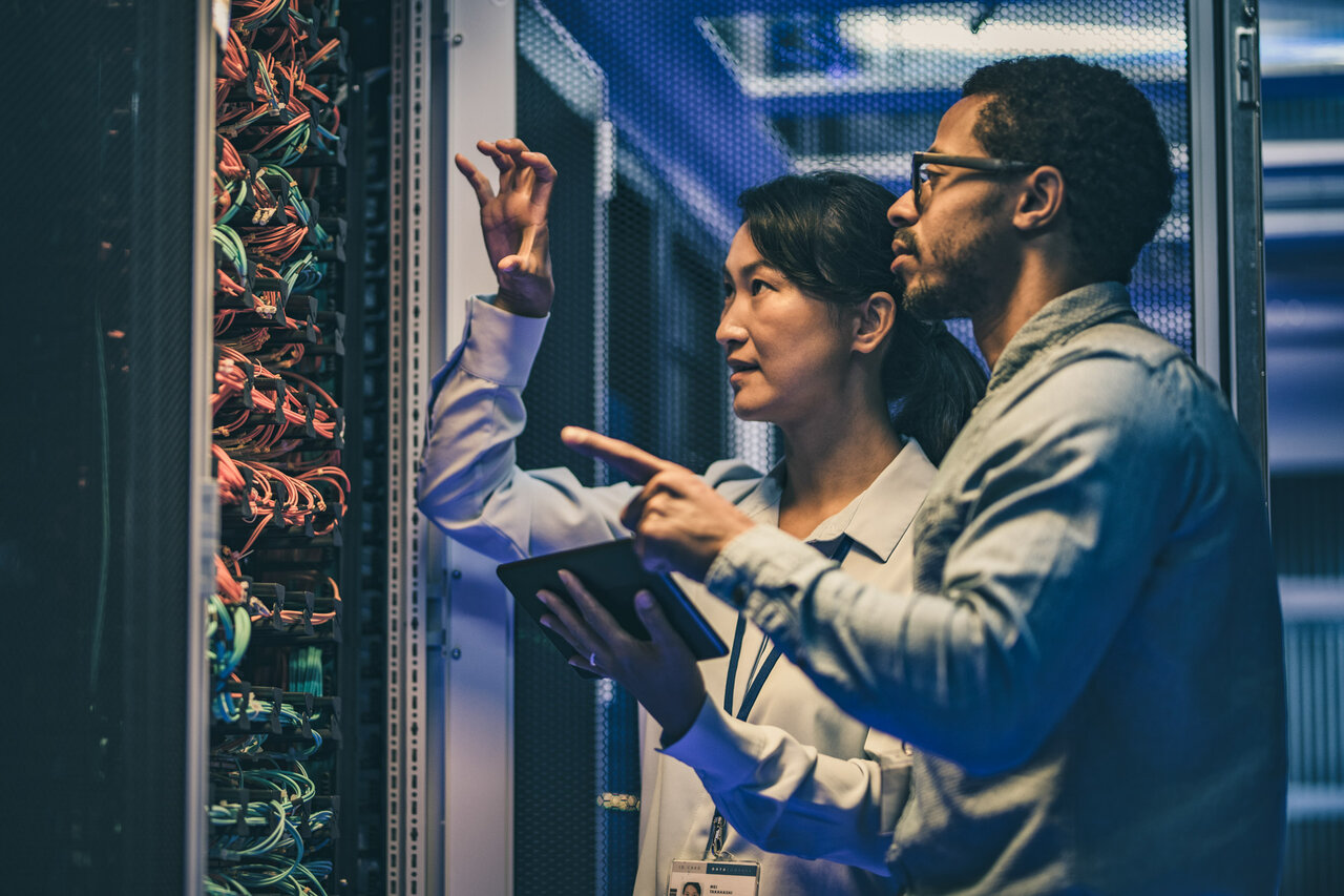 Side view of a female Asian IT engineer explaining network connections in a server to a novice African-American technician. | © vm @getty