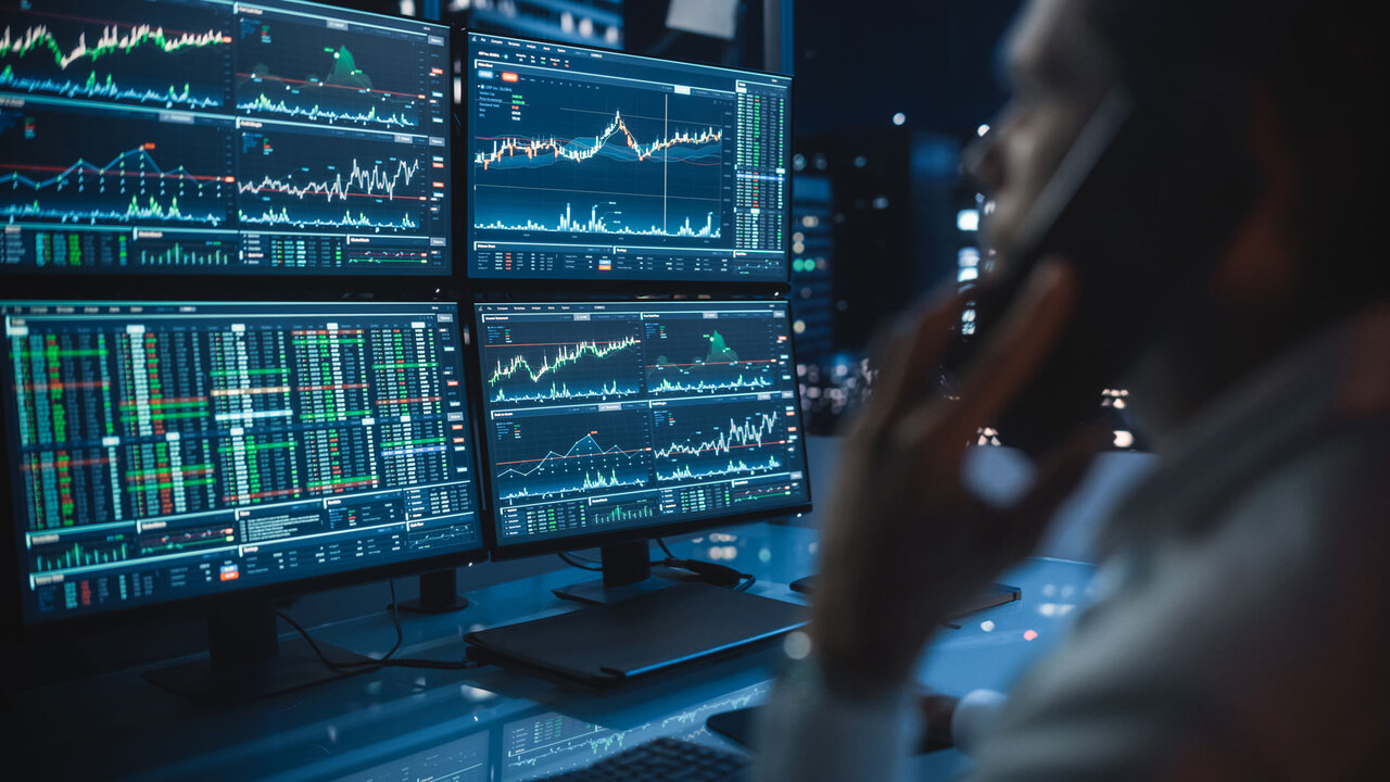 Financial Day Trader Working on a Computer with Multi-Monitor Workstation with Real-Time Stocks Charts and Talking on a Phone with a Client, While in Office Late in the Evening. | © GettyImages