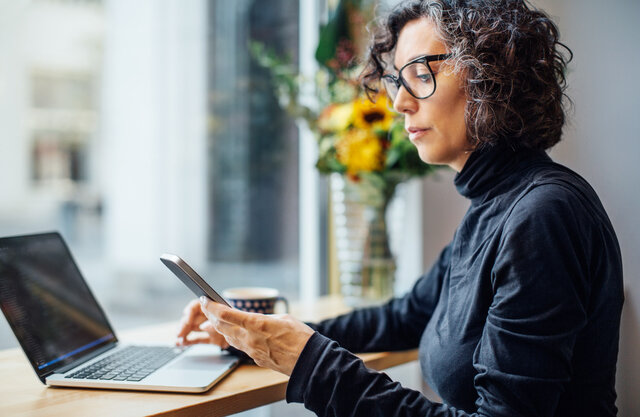 Mature businesswoman sitting at cafe looking at her cell phone while working on laptop computer.  | © Luis Alvarez @GettyImages